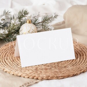 May include: A blank white card stands on a round, woven jute placemat. A sprig of frosted evergreen and a decorative white and gold ornament are in the background. The card is angled, suggesting a place card or invitation.