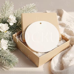May include: A blank, round, white ceramic ornament with a hole for hanging rests inside an open, tan gift box. The box sits on a neutral surface next to a sprig of faux evergreen and white roses. A white, textured fabric is draped nearby.
