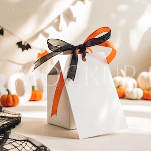 Peut inclure: Une boîte cadeau blanche avec un ruban orange et un nœud noir. La boîte a une étiquette blanche vierge attachée à elle. La boîte est posée sur une surface blanche avec une toile d'araignée noire et blanche au premier plan. Il y a des citrouilles orange et blanches en arrière-plan.