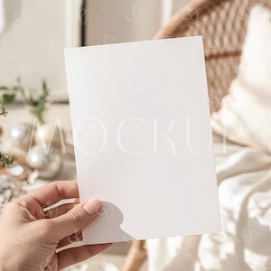 Puede incluir: Una tarjeta rectangular blanca en blanco sostenida en una mano, con un fondo borroso que presenta una silla de mimbre y un cojín blanco. La tarjeta se presenta para resaltar su superficie en blanco, ideal para maquetas o presentaciones de diseño.