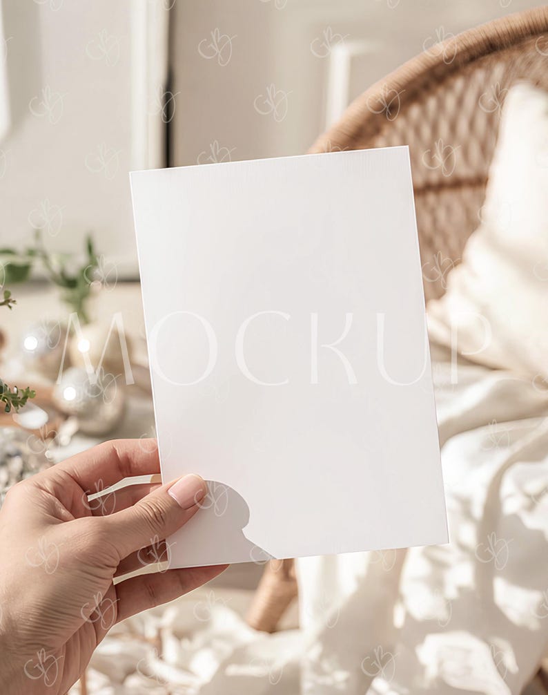 May include: A blank white rectangular card held in a hand, with a soft focus background featuring a wicker chair and a white pillow. The card is positioned to showcase its blank surface, ideal for mockups or design presentations.