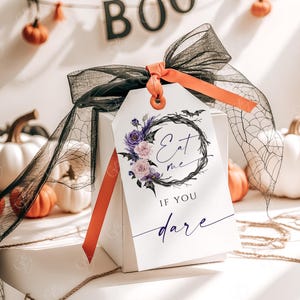 May include: A white gift box with a black and purple floral wreath with bats and the text "Eat me if you dare" on a white tag tied with a black and orange ribbon.