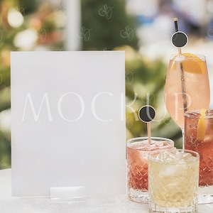 Puede incluir: Un letrero blanco con la palabra "MOCKUP" en una fuente delicada, exhibido sobre un mantel blanco. Varios cócteles en varios vasos de cristal están dispuestos cerca, adornados con rodajas de naranja y palillos de cóctel negros. El fondo está borroso.