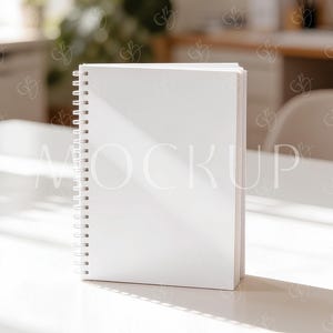 Puede incluir: Un cuaderno blanco con espiral se alza sobre una superficie blanca, bañado por la luz solar. Las páginas del cuaderno están limpias y nítidas, con la palabra "MOCKUP" visible. La escena sugiere una estética limpia y minimalista, ideal para maquetas de papelería o diseño.