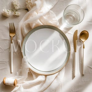 May include: A table setting with a white plate, gold-colored cutlery, and a clear glass. The plate has a light green rim. A white cloth and a sprig of white flowers are also present. The fork, knife, and spoon have white handles.