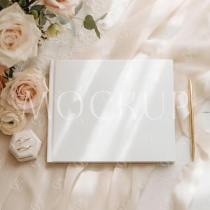 May include: A white book with the word "MOCKUP" faintly visible, next to a gold pen and a ring box with two rings. The scene is set on a cream-coloured fabric with a bouquet of peach roses.