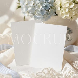 May include: Two blank white cards with the word "MOCKUP" faintly visible, resting on a cream-colored surface with lace and a blue and white striped ribbon. A vase of blue and white hydrangeas is in the background.