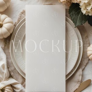 May include: A styled table setting with a blank menu card. The scene includes white pumpkins, a floral arrangement, and a gold knife. The plates and linen napkin add to the elegant, neutral color palette. The word "MOCKUP" is faintly visible on the card.