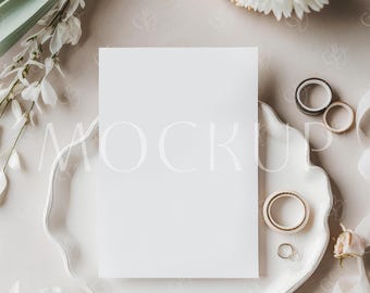 Può includere: Un biglietto bianco vuoto è posto su un piatto bianco con bordo smerlato, circondato da accessori per matrimoni. La scena comprende anelli d'oro, nastro adesivo decorativo e fiori bianchi, creando un'estetica elegante e minimalista.