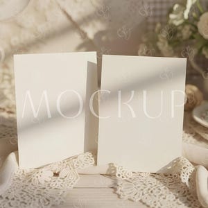Op de afbeelding: Twee blanco, crèmekleurige vierkante kaarten met het woord "MOCKUP" erop gedrukt, op een wit houten oppervlak. De kaarten worden omringd door kant en zacht licht, ideaal voor een mockup van briefpapier of uitnodigingen.