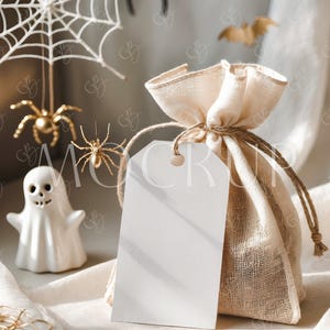 Könnte beinhalten: Ein weißer Geschenkanhänger mit einem leeren Feld zum Beschreiben ist an einem kleinen, beigefarbenen Jutesack mit Kordelzug befestigt. Der Sack liegt auf einer weißen Stoffoberfläche mit einer weißen Geisterfigur und goldenen Spinnendekorationen im Hintergrund.