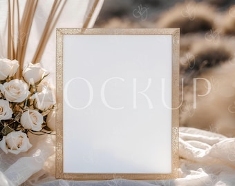 May include: A gold rectangular frame with an ornate border, displaying the word "MOCKUP" in a soft, light font. The frame rests on a white cloth, with a bouquet of white roses and dried plants in the background. The scene is set outdoors.