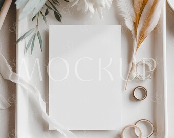 Può includere: Una carta rettangolare bianca con la parola "MOCK" in grigio chiaro, posta su un vassoio bianco. Il vassoio è decorato con fiori bianchi, foglie verdi, piume e anelli. Sono presenti anche un nastro bianco e un nastro beige.