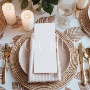 Op de afbeelding: Een witte kaart met een lege ruimte voor tekst ligt op een beige gestreepte servet. De servet is gevouwen en ligt op een geweven placemat. De placemat ligt op een wit tafelkleed met een bruin bladmotief. Er staan twee witte kaarsen en een wijnglas op de tafel.