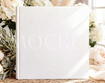 May include: A white book with the word "MOCKUP" faintly visible, resting on a woven surface. The book is surrounded by white flowers and greenery, creating a soft, natural aesthetic. The scene is illuminated by natural light.