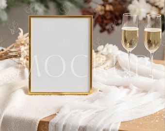 May include: A gold framed picture frame with a white interior sits on a wooden table. Two champagne flutes filled with a bubbly beverage are to the right. The table is covered with a white cloth and decorated with dried flowers.