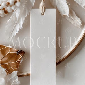 May include: A blank, rectangular white tag with a hole at the top, suspended by beige twine. The word "MOCKUP" is faintly visible on the tag. The tag is surrounded by white feathers, dried leaves, and a wooden tray.