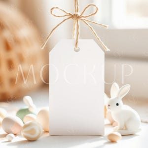 May include: A white gift tag with a twine bow hanging from it. The tag is blank and ready for a message. The tag is surrounded by pastel colored Easter eggs and a white bunny figurine.