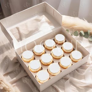 May include: A dozen cupcakes in a white box with a clear window. Each cupcake has a white circular top and a golden-brown base. The box is on a beige fabric surface, with decorative elements in the background.