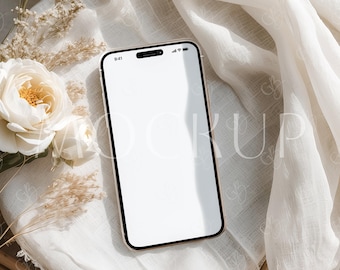 May include: A white smartphone with a blank screen is lying on a white fabric surface with dried flowers and a woven basket underneath.