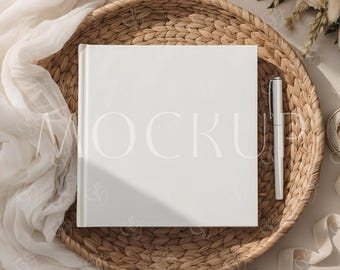 May include: A white square book with the word "MOCKUP" printed on the cover, placed on a woven tray. A silver pen rests beside the book. The scene includes white fabric, flowers, and ribbons, creating a soft, elegant aesthetic.