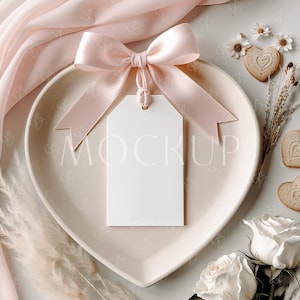 Könnte beinhalten: Ein weißer leerer Geschenkanhänger mit einem rosa Satinband, das zu einer Schleife gebunden ist, auf einem herzförmigen Teller. Der Teller befindet sich auf einer weißen Oberfläche mit getrockneten Blumen und weißen Rosen.
