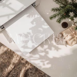 May include: A flat lay featuring a white printer with a blank white paper, a faux fur blanket, a wrapped gift, pine branches, a pine cone, and decorative snowflakes. The scene is bathed in natural light, creating shadows.