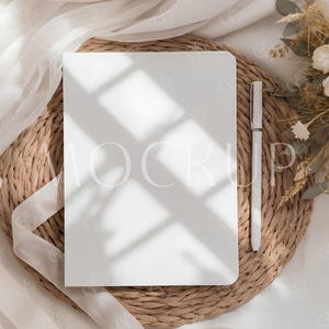 Puede incluir: Un cuaderno blanco con la palabra "MOCKUP" visible y un bolígrafo plateado descansan sobre un salvamanteles tejido. También hay una cinta blanca y flores secas. La escena está iluminada con luz natural suave, creando sombras.