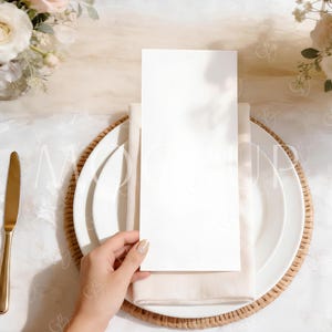 May include: A blank white menu card is held over a white plate, napkin, and woven charger. A gold knife is on the left. The table is decorated with flowers and a cream-colored tablecloth. The scene is set for a wedding or special event.