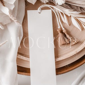 Op de afbeelding: Een blanco, rechthoekig wit label met een touwtje en twee beige kwastjes hangt over een houten kom. Het label is het middelpunt, met gedroogd wit en beige blad en een witte doek op de achtergrond. De scène heeft een neutrale, minimalistische esthetiek.