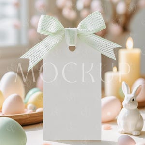 May include: A white gift tag with a green bow tied around it. The tag is sitting on a table with pastel colored Easter eggs, a white ceramic bunny, and a candle in the background.