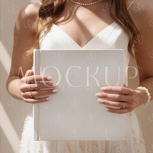 May include: A bride wearing a white wedding dress holds a white photo album in front of her. The album has a blank cover.