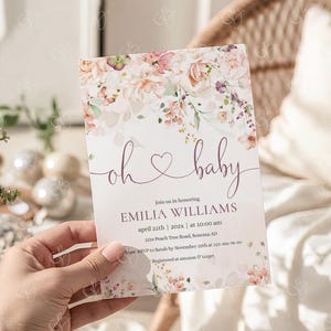 May include: A baby shower invitation with floral accents in pink and peach tones. The text "oh baby" is written in a delicate script, with event details below. The invitation is held in a hand.