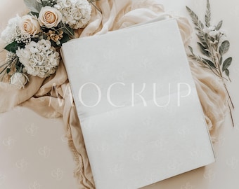 May include: A white blank card mockup with the word "MOCKUP" printed in white on the card. The card is lying on a beige fabric with white flowers and green leaves.