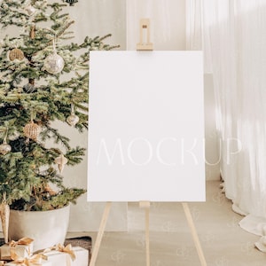 Può includere: Una tela bianca su un cavalletto con una cornice in legno. La tela è vuota e pronta per un design. Il cavalletto è di fronte a un albero di Natale con ornamenti dorati.