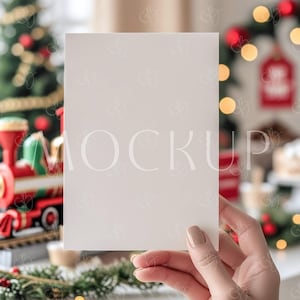 以下が含まれることがあります： 手で持たれた白い無地のカード。背景にはクリスマスツリーとお祝いの飾り付け。カードには「MOCKUP」の文字が薄く表示されています。このシーンはホリデーシーズンを連想させます。