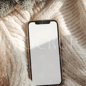 May include: A gold-framed smartphone with a blank white screen rests on a cream-colored, chunky knit blanket. The blanket is draped, creating soft folds and shadows. A sprig of frosted greenery is in the upper left corner, adding a touch of wintery decor.