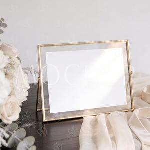 May include: A gold-framed glass picture frame with a white insert stands on a dark wooden surface. A bouquet of white and cream-colored flowers and a cream-colored fabric are in the background. The frame is angled, and the glass reflects light.