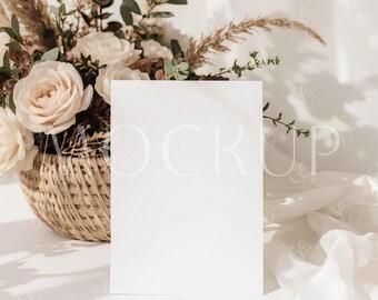 Può includere: Una carta bianca vuota è posizionata al centro. Un cesto di vimini traboccante di rose color crema ed erbe secche è a sinistra. Lo sfondo presenta una luce soffusa e un drappo di tessuto bianco, creando un'estetica minimalista ed elegante.