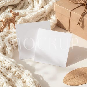 May include: A styled product mockup featuring two white square cards with the word "MOCKUP" faintly visible. A beige gift box tied with twine, a gold deer figurine, a gold leaf, and a cream-colored knitted scarf complete the scene.