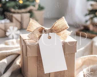 May include: A wrapped gift box with a blank white tag, tied with a burlap ribbon and bow. The box is wrapped in tan paper and sits on a soft, cream-colored blanket. A Christmas tree is visible in the background.