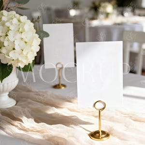 May include: A wedding table setting with a white floral arrangement in a white vase, blank white cards in gold holders, and a beige fabric runner. The scene is bathed in natural light, creating a soft, elegant ambiance.