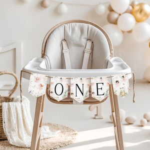 May include: A white high chair with a floral banner that says "ONE" hanging from it. The banner has a pink, green, and white floral pattern with a small fairy illustration on each flag.