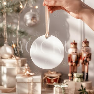 May include: A plain, white, circular Christmas bauble with a gold ribbon, held up by a hand. The background includes a Christmas tree, wrapped presents, and nutcrackers, creating a festive holiday scene.