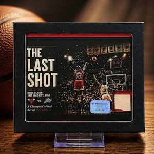 May include: A framed sports collectible titled The Last Shot featuring Michael Jordan in a red jersey, a basketball court scene, the text June 14 1998 Delta Center Salt Lake City Utah, a Bulls and Jazz logo, and a small wood piece with a holographic sticker.