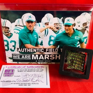 May include: A certificate of authenticity for a We Are Marshall movie prop. The certificate is white with black text and a black and white image of a football player. The certificate has a serial number of 243756. The prop is a small, green, square-shaped object in a black frame.