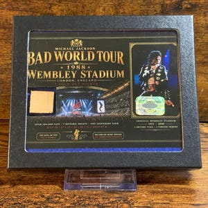 May include: A framed collectible featuring Michael Jackson's Bad World Tour at Wembley Stadium in 1988. The image includes a photo of Jackson performing, the tour logo, and text indicating sold-out nights. The frame is black.