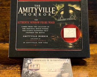 Könnte beinhalten: Eine schwarze Vitrine mit einem kleinen, quadratischen Stück weißem, rissigem Holz, das als authentisches Fensterrahmenholz aus dem Amityville Horror-Haus gekennzeichnet ist. Die Box enthält Text zum Standort und ein Echtheitszertifikat.