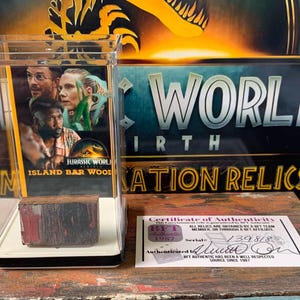 Könnte beinhalten: Ein kleiner Holzblock, rot und schwarz bemalt, wird in einem durchsichtigen Acryl-Würfel ausgestellt. Der Würfel steht auf einem weißen Sockel. Der Block ist eine Requisite aus dem Film "Jurassic World: Fallen Kingdom". Im Hintergrund ist ein Echtheitszertifikat zu sehen, mit dem Text "Echtheitszertifikat" und "BET Authentic ist seit 1987 eine seriöse und angesehene Quelle".