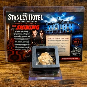 Könnte beinhalten: Eine rechteckige Vitrine mit einem rauen, beigefarbenen architektonischen Steinfragment. Dahinter befindet sich eine Karte mit dem Stanley Hotel, dem Text "Filming Location of The Shining" und einem Echtheitszertifikat.